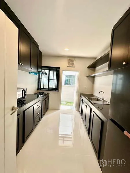 Detached House for rent in Bang Phli, Samut Prakan - Galley kitchen with black cabinetry, gas hob, sink and window, leading to the rear yard.