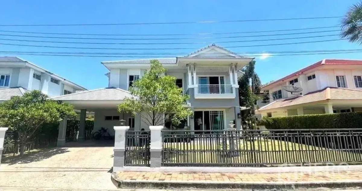 Detached House for sale in Samut Prakan - Street view of a gated two-storey house with lawn and trees in an upscale community.