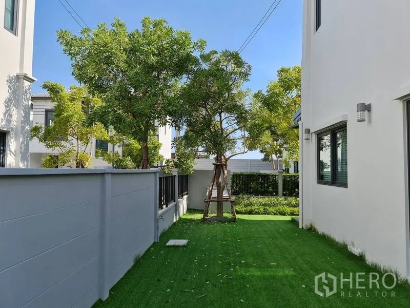Detached House for rent in Samut Prakan - Side garden with artificial grass lawn, shade trees and privacy wall.