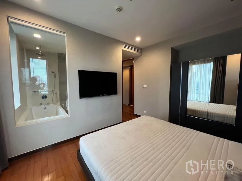 Condominium for rent - Master bedroom with wall‑mounted TV and glass window looking into an ensuite bathroom with bathtub.