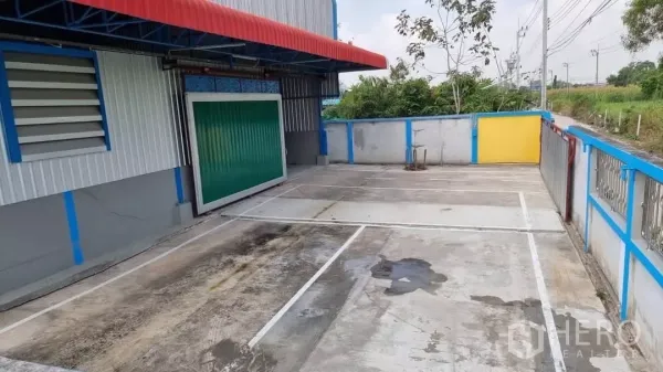 货栈 出租在Min Buri, Bangkok - 私家混凝土庭院，划线停车位，绿色卷帘装卸门与遮雨棚。