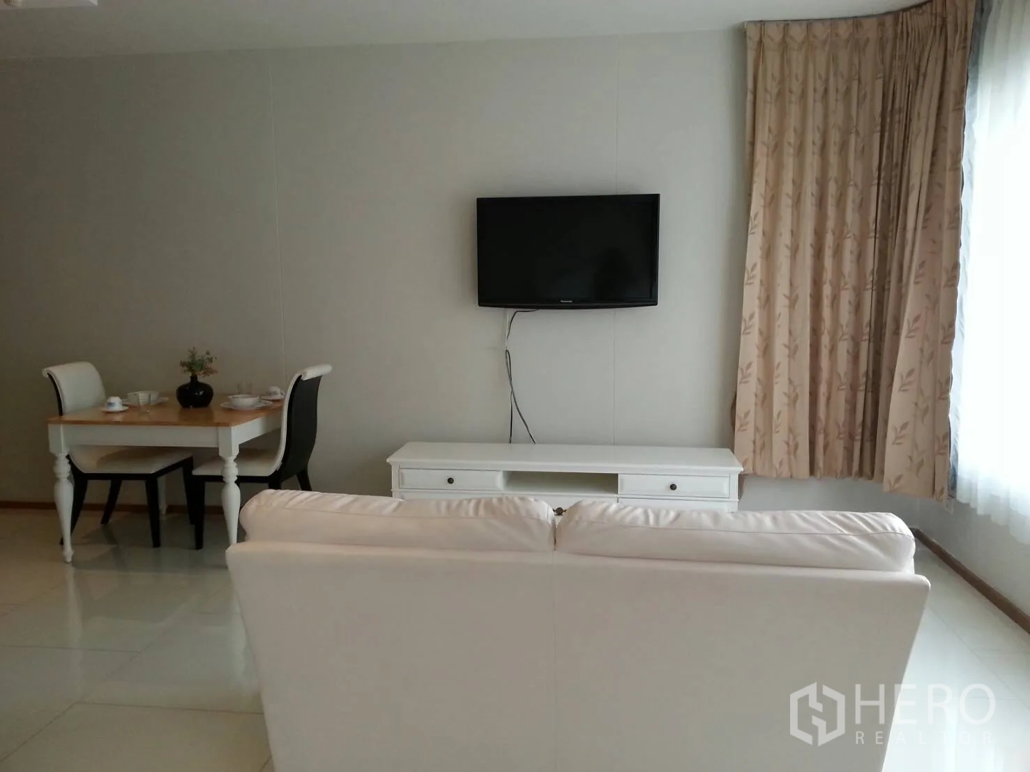 Condominium for rent in Khlong Toei, Bangkok - Living room with TV console and two-seat dining table next to full-height curtained windows.