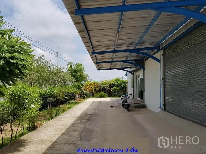 Factory for rent in Samut Sakhon - Covered front apron and driveway beside landscaped greenery and shutter door.