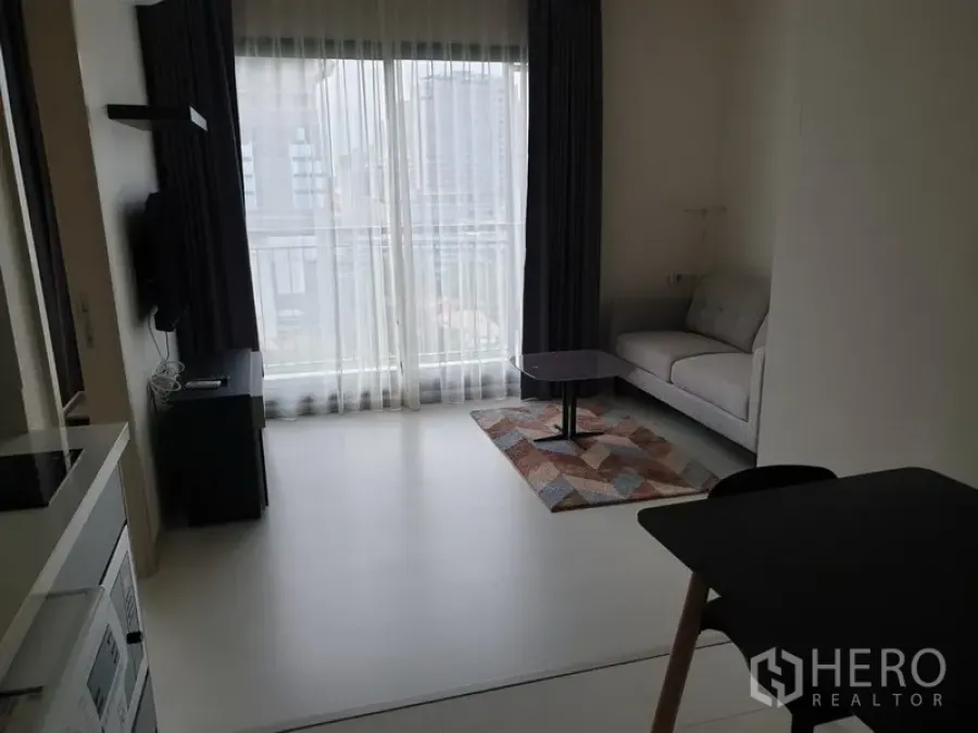 Condominium for rent in Khlong Toei, Bangkok - Bright living room with floor-to-ceiling window, sheer curtains, grey sofa and small coffee table, offering a city view in a Khlong Toei condo near BTS Thonglor.