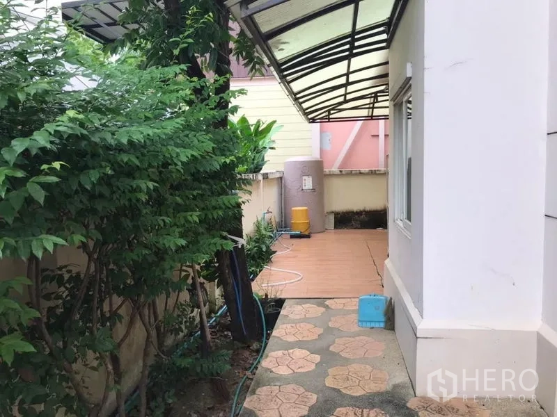 Detached House for rent in Bang Khen, Bangkok - Shaded side path with canopy roof, utility tank and paved area amid greenery.