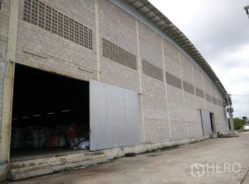 Factory for rent or sale in Krathum Baen, Samut Sakhon - Long warehouse facade with sliding metal doors and a wide concrete apron for trucks.