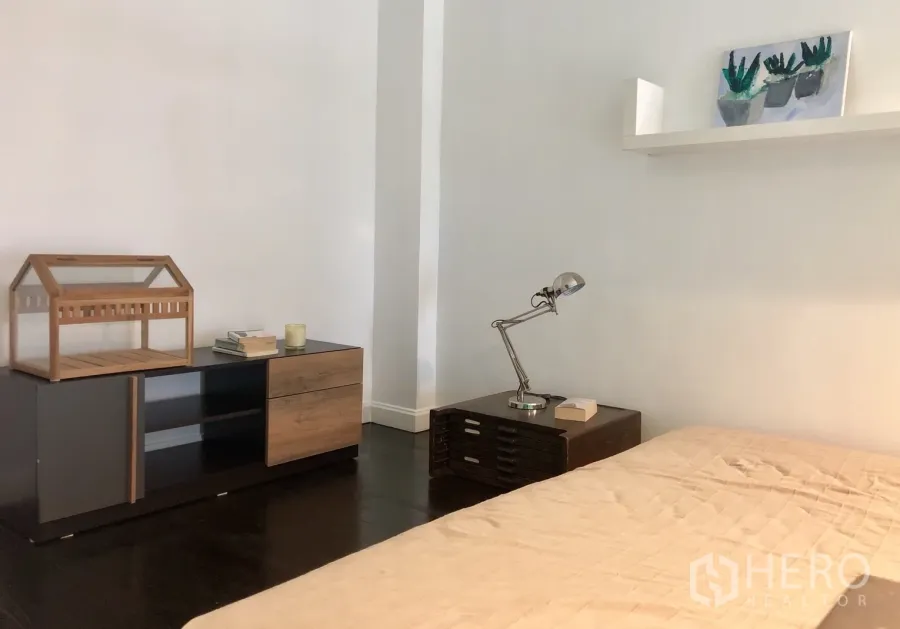 Condominium for rent or sale in Khlong Toei, Bangkok - Minimalist bedroom with dark wood floor, bedside lamp and a low cabinet with decor.
