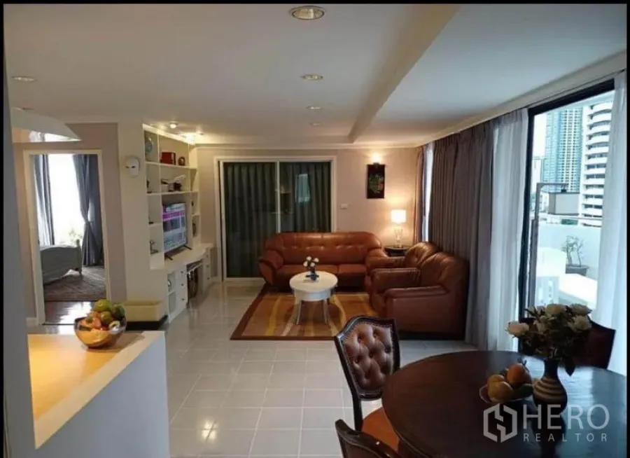 Condominium for rent or sale in Khlong Toei, Bangkok - Bright Sukhumvit condo living room with brown leather sofas, built‑in shelves and sliding doors to a city‑view balcony.