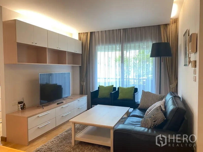 Condominium for rent or sale in Khlong Toei, Bangkok - Bright living room with TV wall unit, leather sofa and sheer curtains opening to a balcony.
